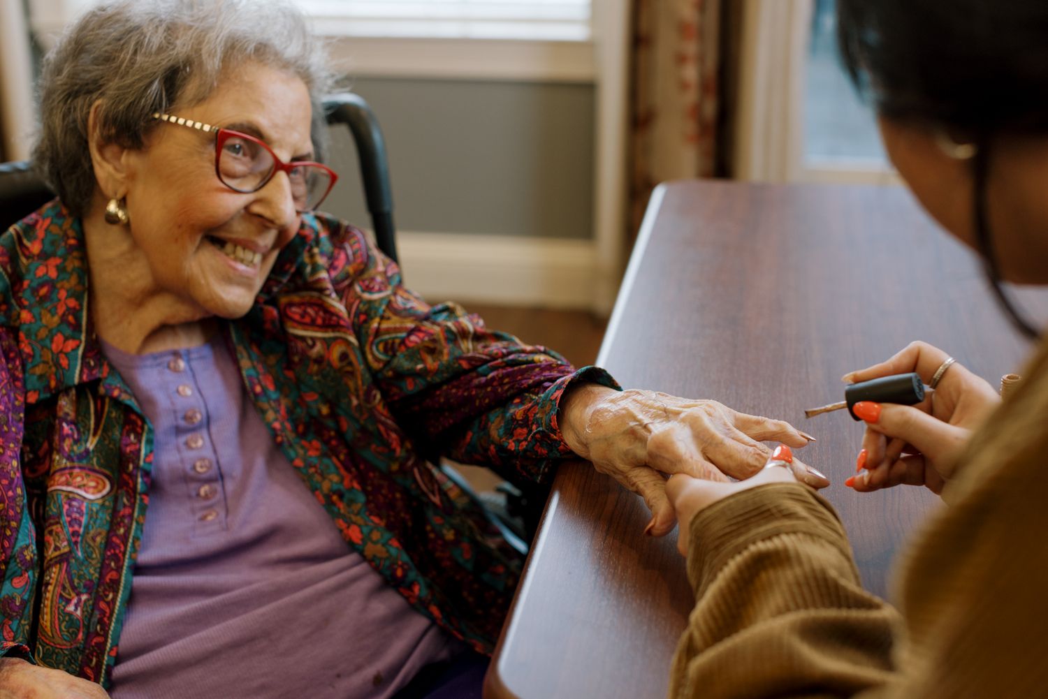 Elderly woman smiling joyfully while getting her nails painted by a caregiver at a senior care facility.