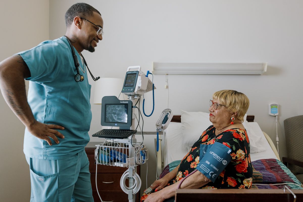 Male nurse in scrubs checking blood pressure of elderly female patient in hospital room.