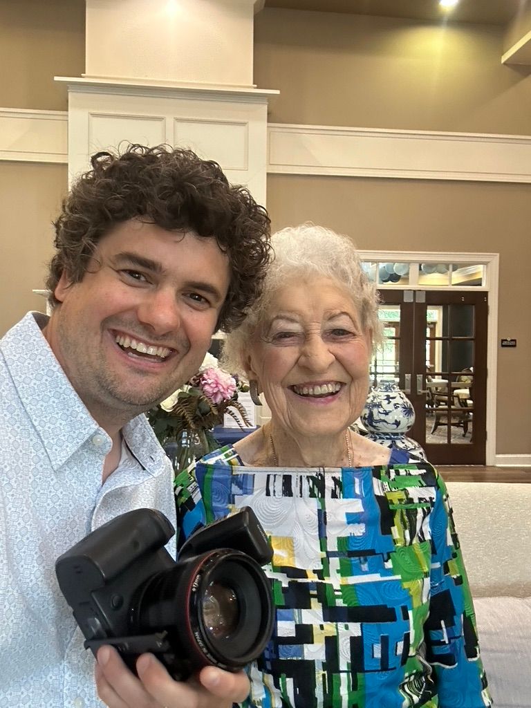 Smiling photographer takes a selfie with an elderly woman holding a wide-angle camera lens.