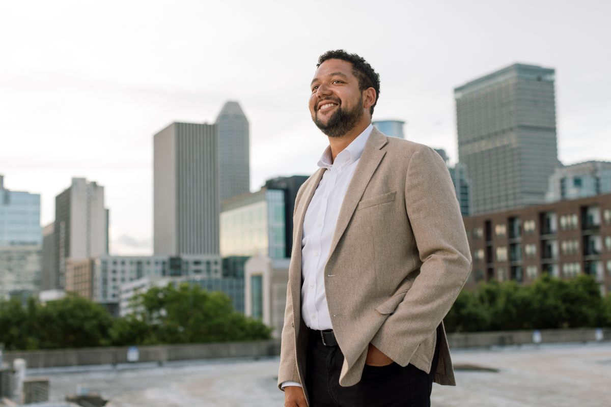 Smiling businessman in beige blazer stands confidently outdoors with city skyline in background.
