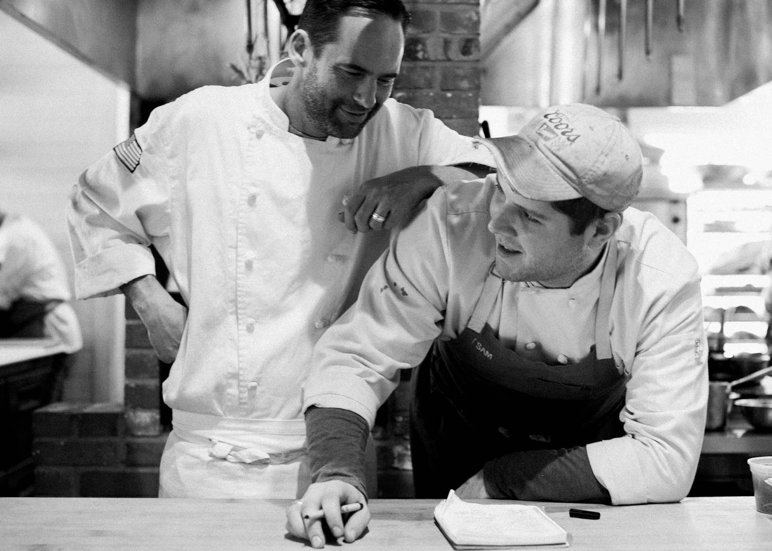 Two chefs in white uniforms share a laugh while reviewing notes in a professional kitchen.