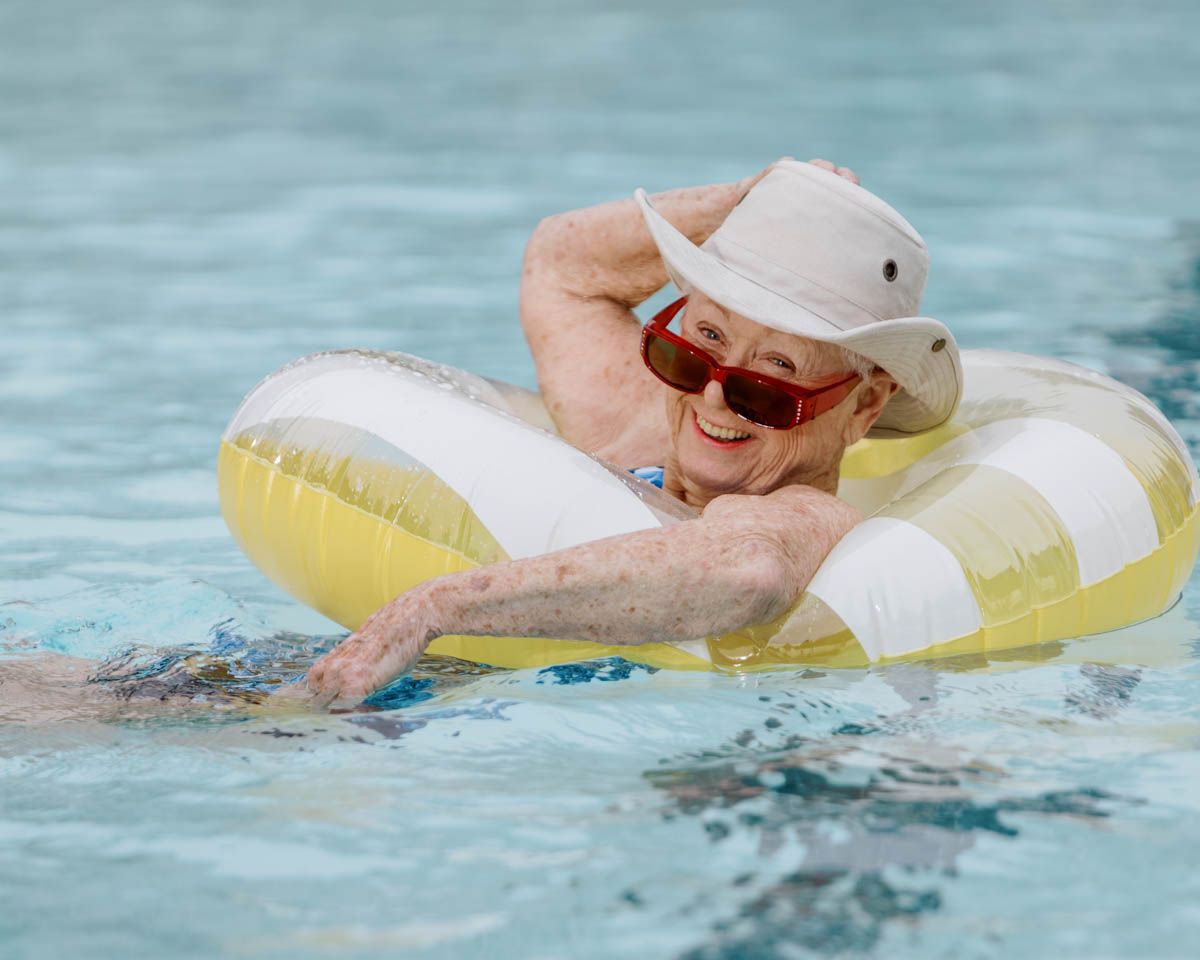 Happy senior woman relaxing on inflatable ring in pool, wearing white hat and red sunglasses.