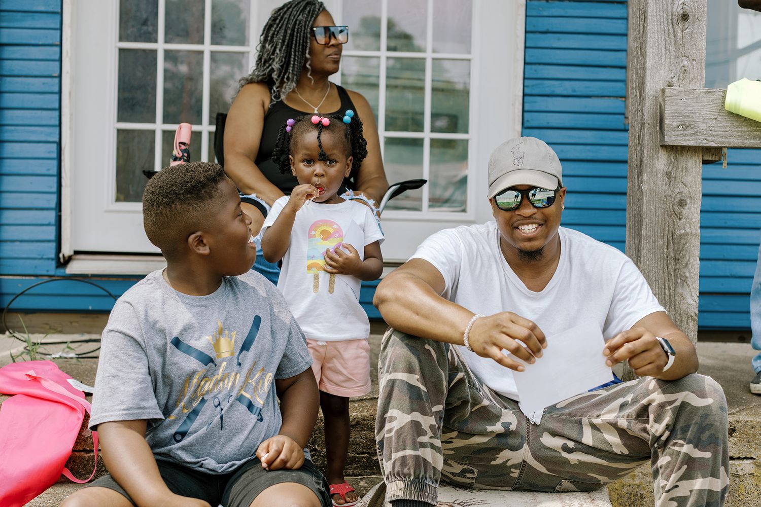 Happy Black family relaxing together on steps outside a blue house on a sunny day.