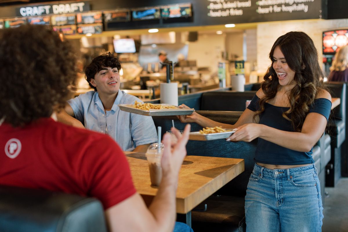 Smiling waitress serves pizza to customers at a casual restaurant with craft draft beer signs.