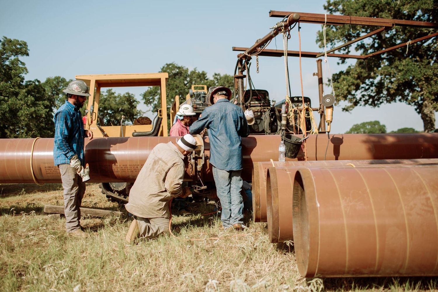Workers in hard hats assemble large steel pipeline sections in a field using heavy machinery.