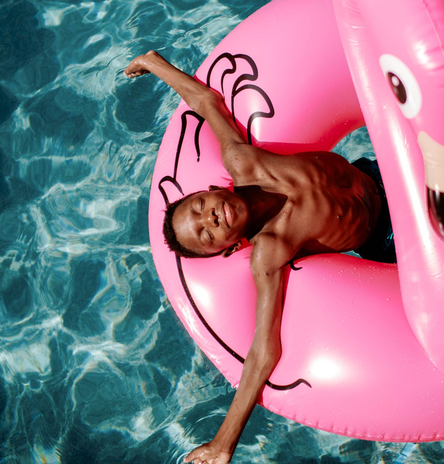 Shirtless boy relaxing in a pink flamingo inflatable pool float in a sunny swimming pool.