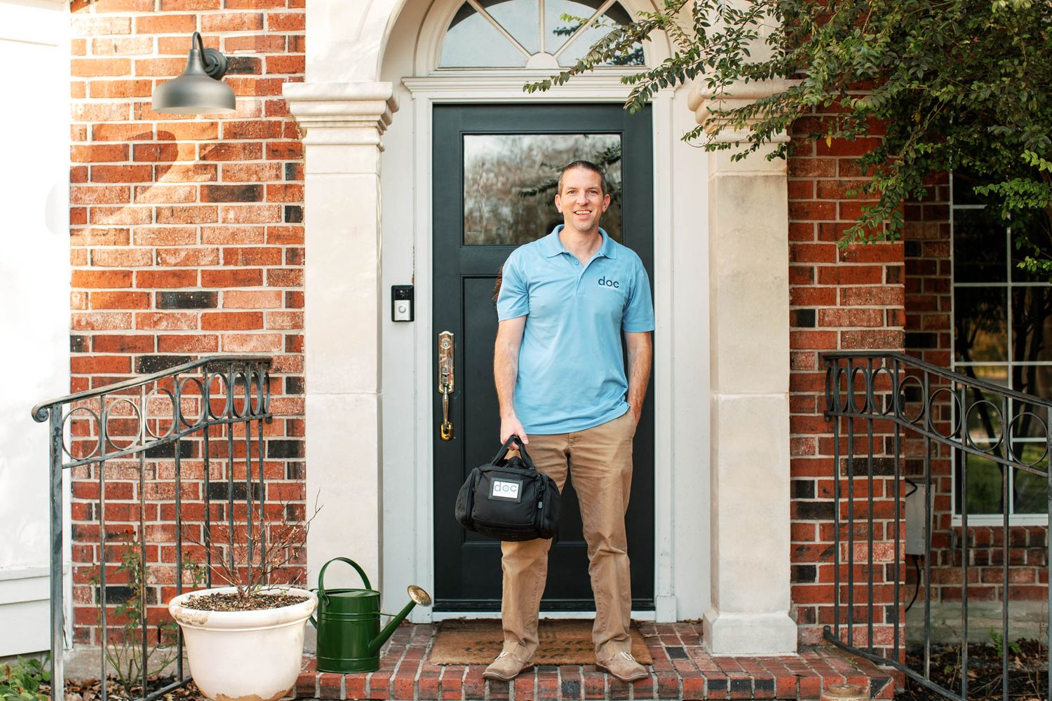 A healthcare professional in a blue polo stands at a brick home's front door holding a medical bag.