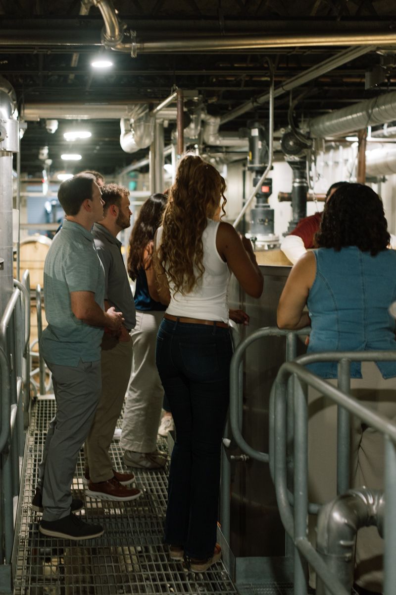 Group of people on an industrial facility tour, standing on metal grating surrounded by pipes and machinery.