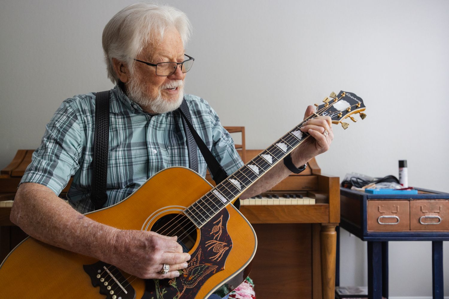 Elderly man with white beard playing acoustic guitar, wearing plaid shirt and suspenders, with piano in background.
