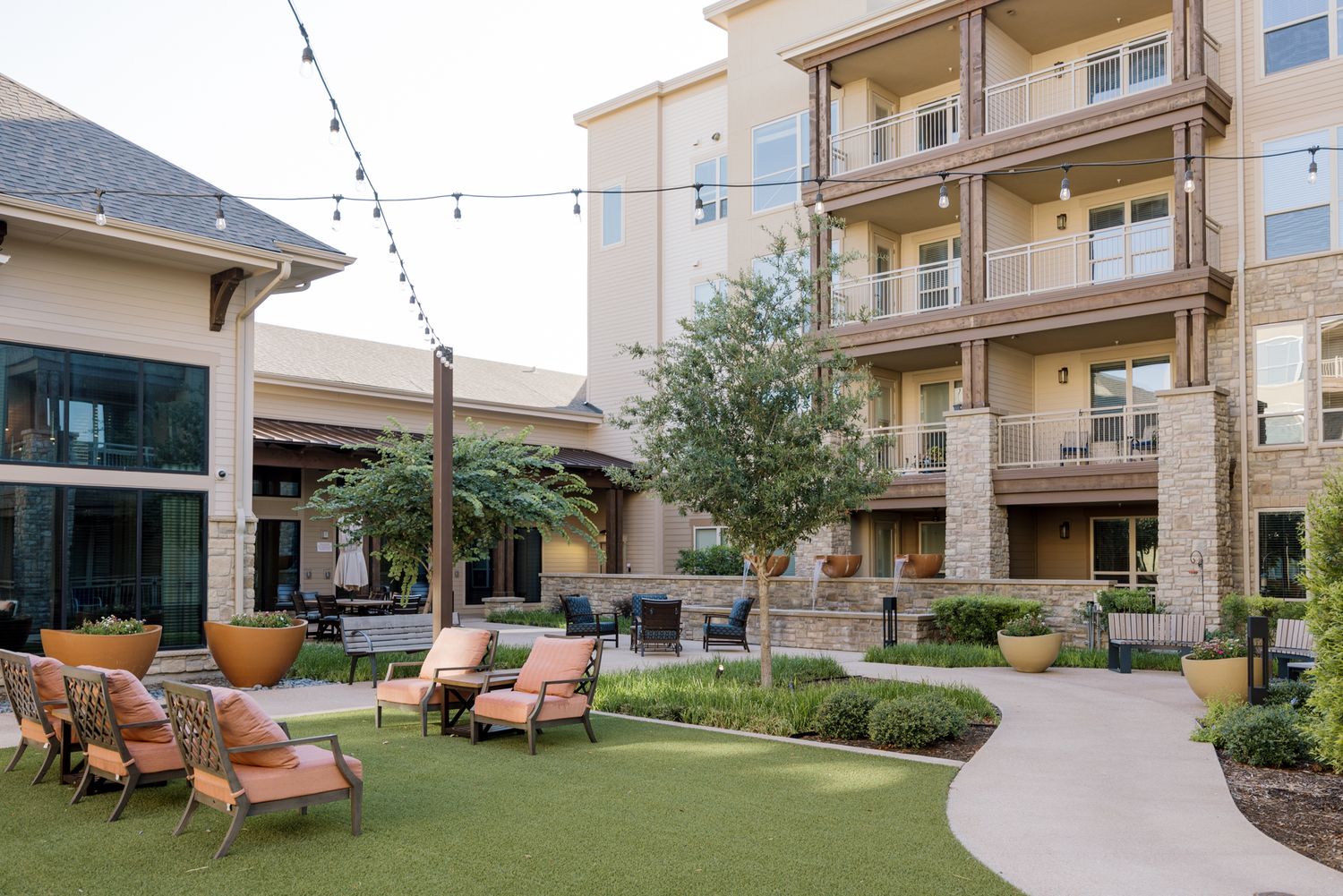 Luxury apartment courtyard with string lights, lounge seating, manicured lawn, and multi-story balconies.