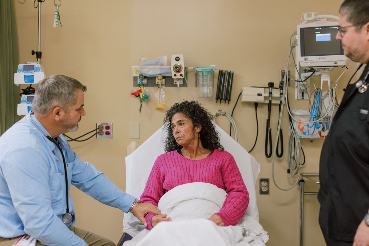 Doctor consulting with female patient lying in hospital bed while medical staff assists nearby.