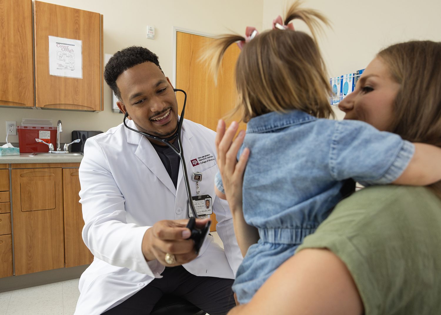 A smiling medical professional in a white coat examines a young child with a stethoscope.