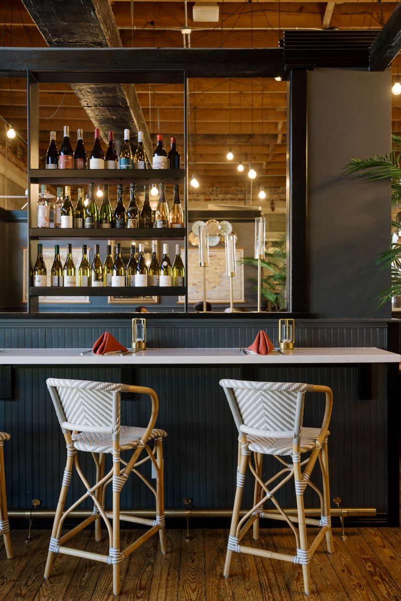 Rustic bar with rattan stools, white marble counter, red napkins, and wine bottle shelves lit by Edison bulbs.