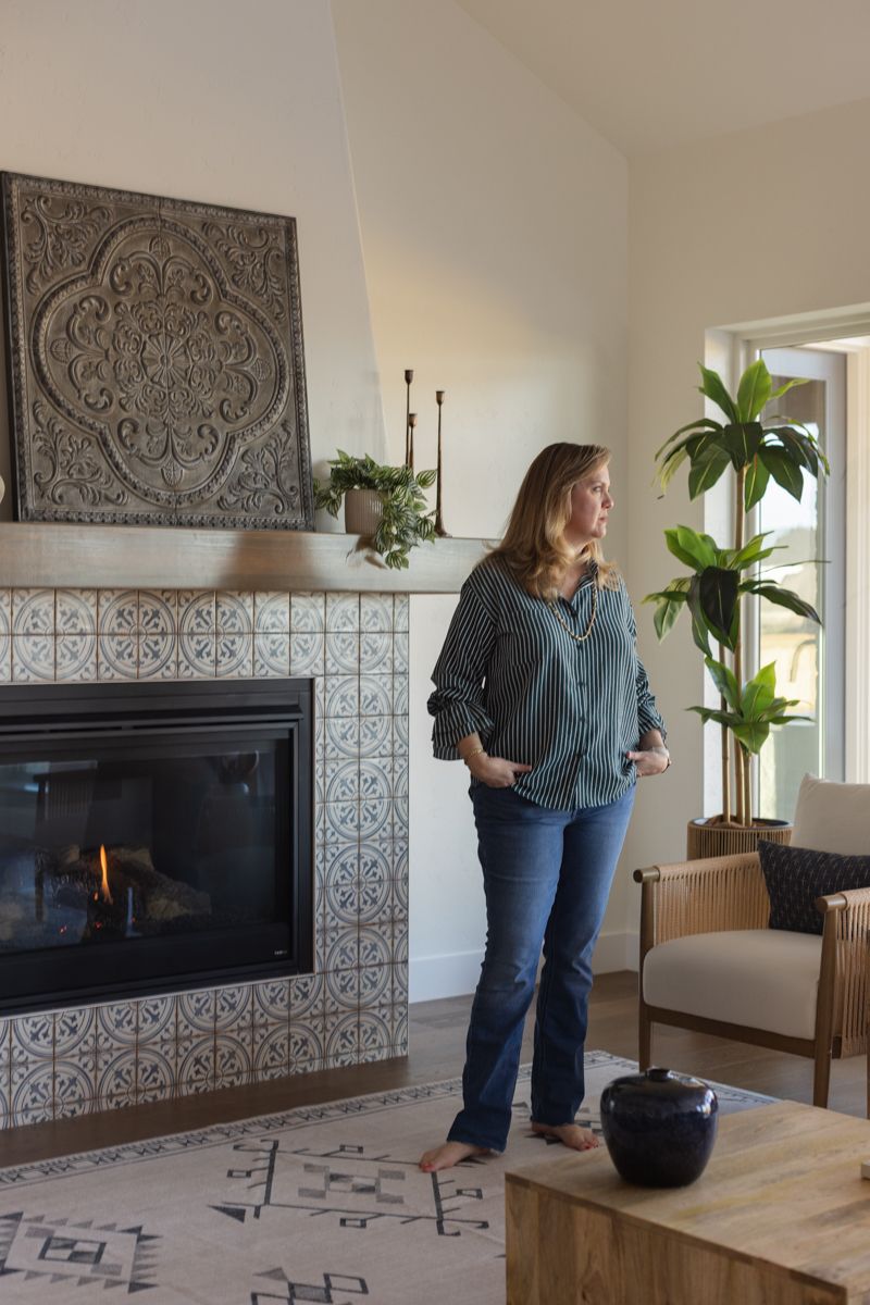 Woman standing in bright living room with decorative tile fireplace, ornate wall art, and lush indoor plants by window.