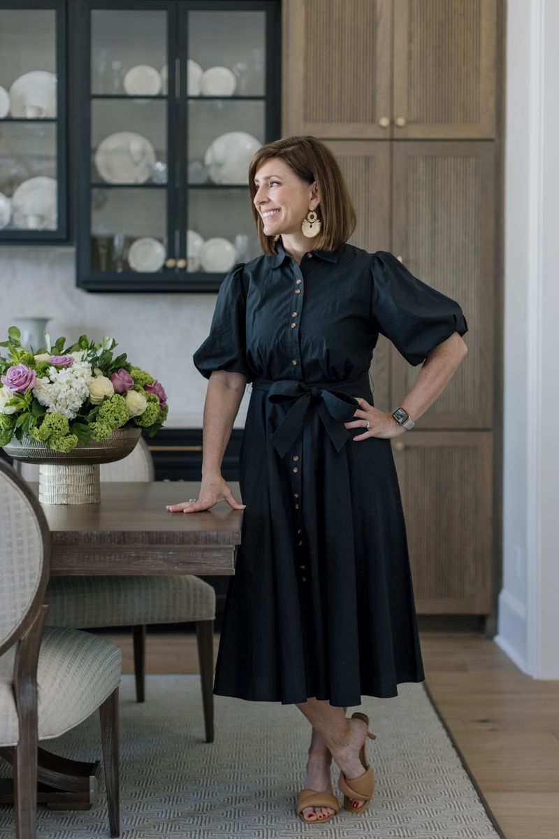 Woman in black midi dress posing in elegant dining room with wood furniture and glass-front china cabinet.