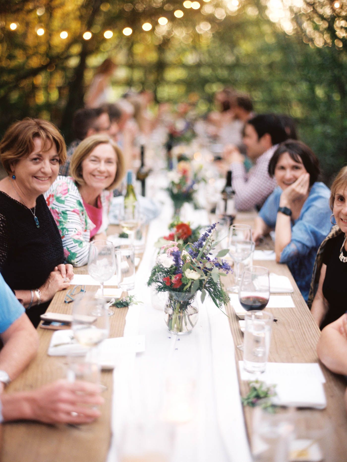 Guests enjoying an outdoor dinner party at a long wooden table with floral centerpieces and string lights.