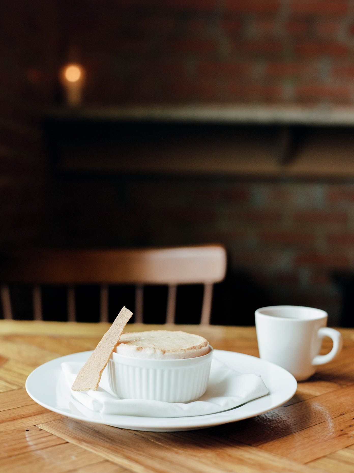 A warm souffle in a white ramekin served on a plate with a wooden spoon and espresso cup in a cozy restaurant.