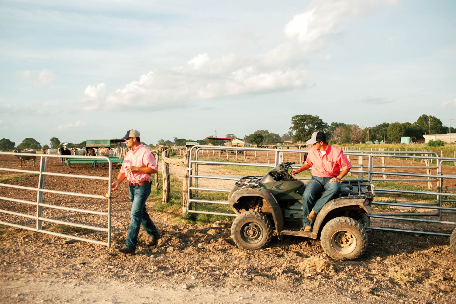 Two farmers on a ranch, one sitting on an ATV near metal cattle gates on a sunny day.