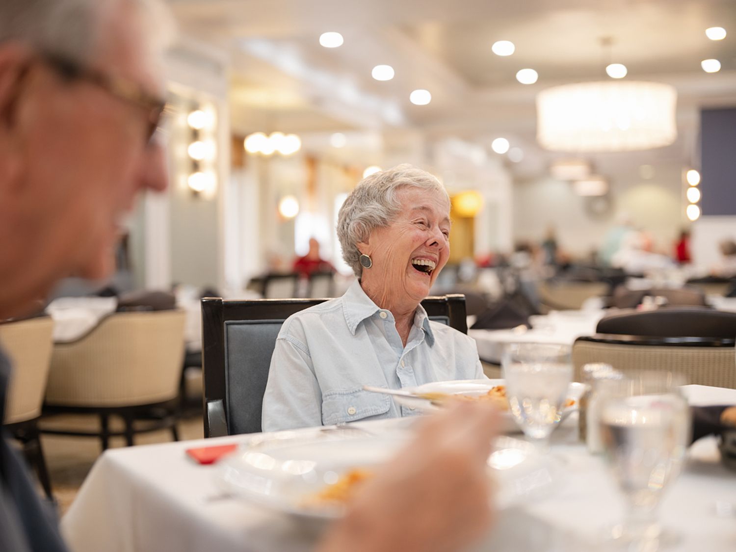 Elderly woman laughing joyfully while dining with a companion in an elegant restaurant setting.