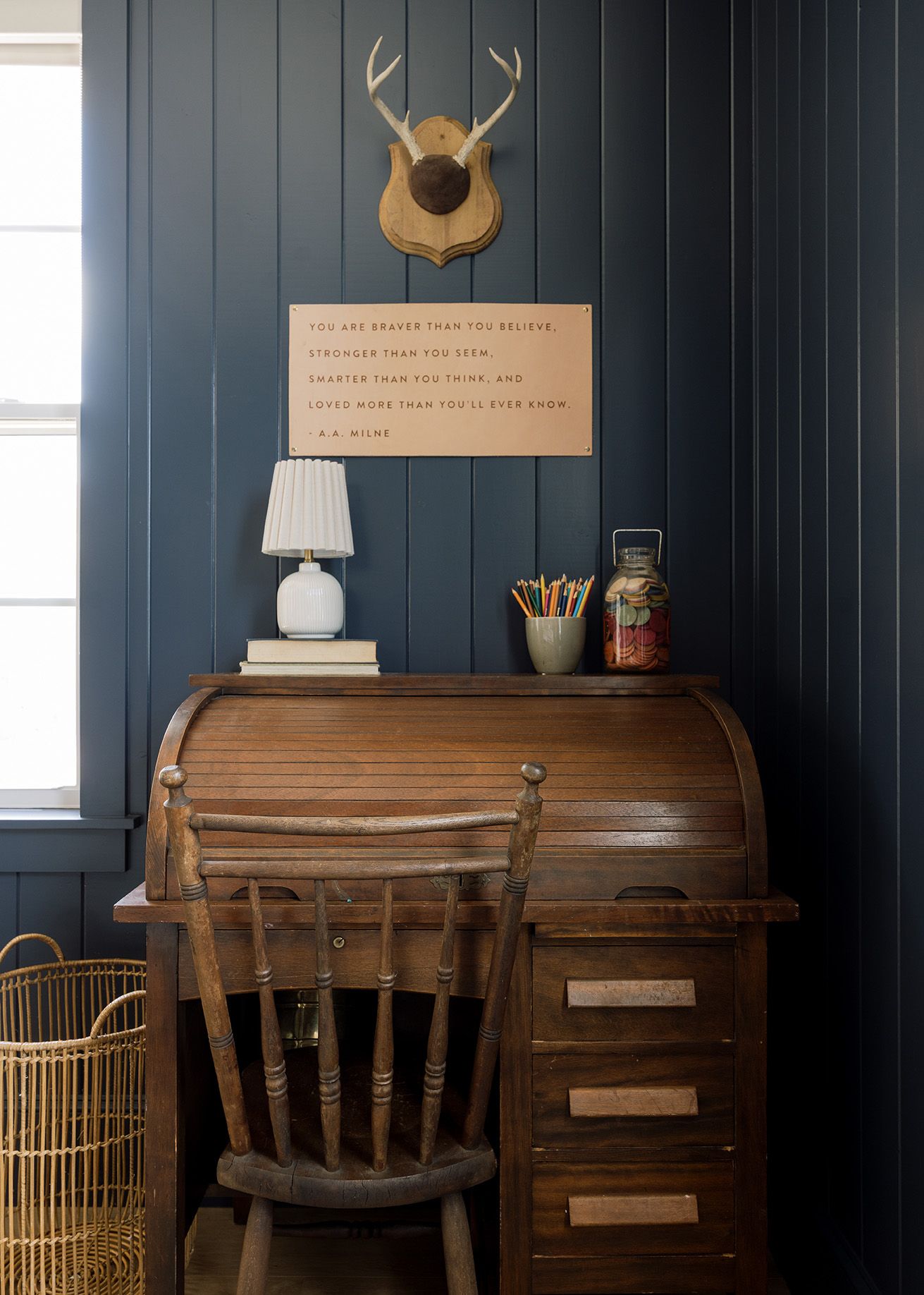 Vintage roll-top desk with wooden chair against dark navy shiplap wall with antler mount decor.