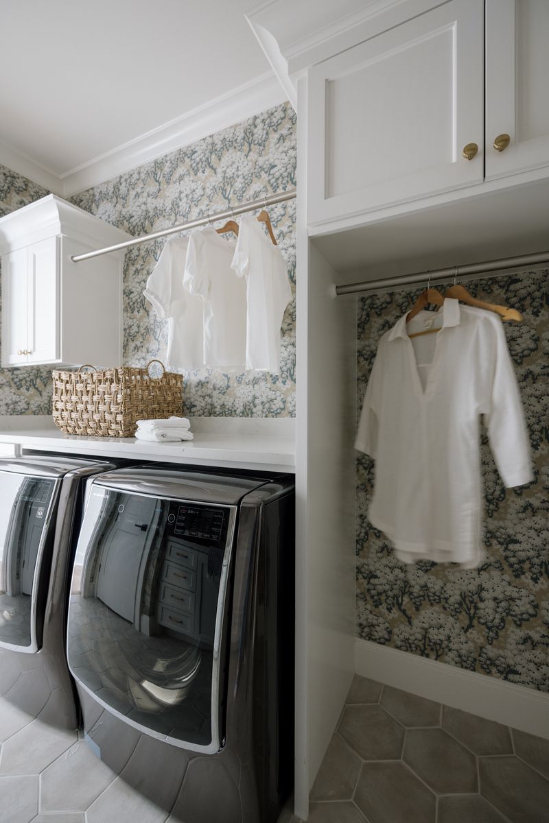 Modern laundry room with floral wallpaper, white cabinets, stainless appliances, and hanging white shirts on a rod.