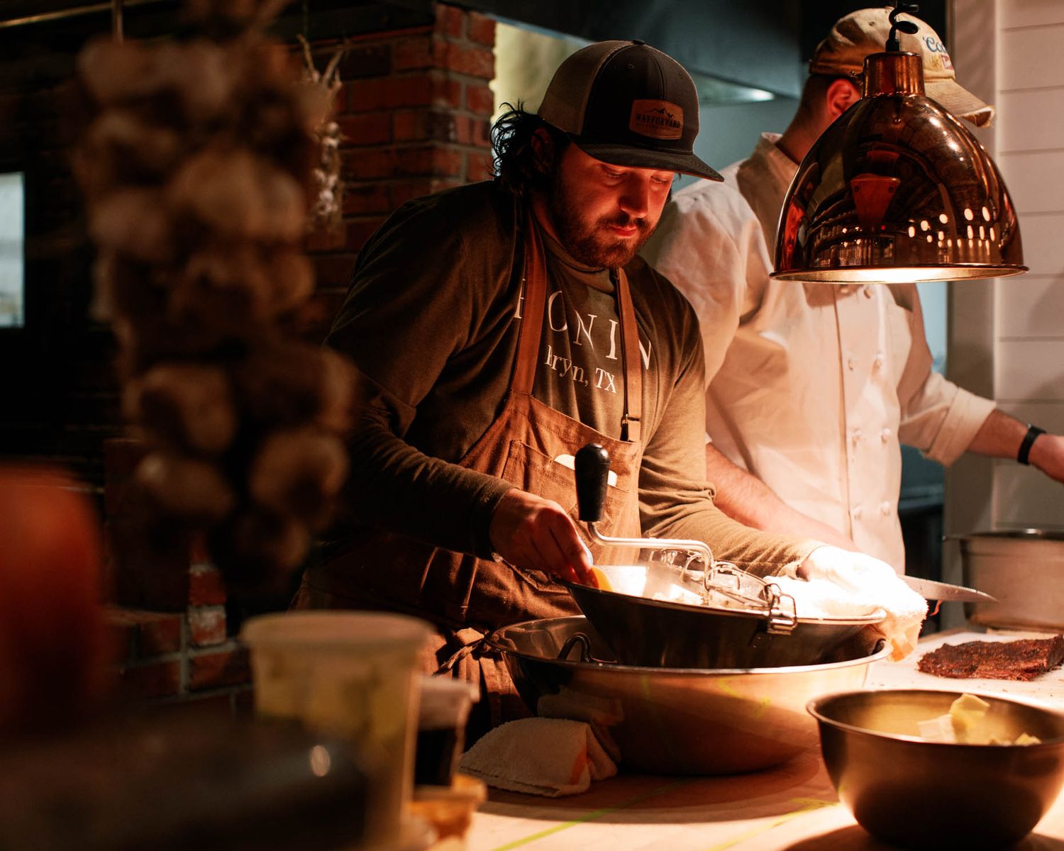 Chef cooking in a restaurant kitchen under warm pendant lights with garlic hanging in background.