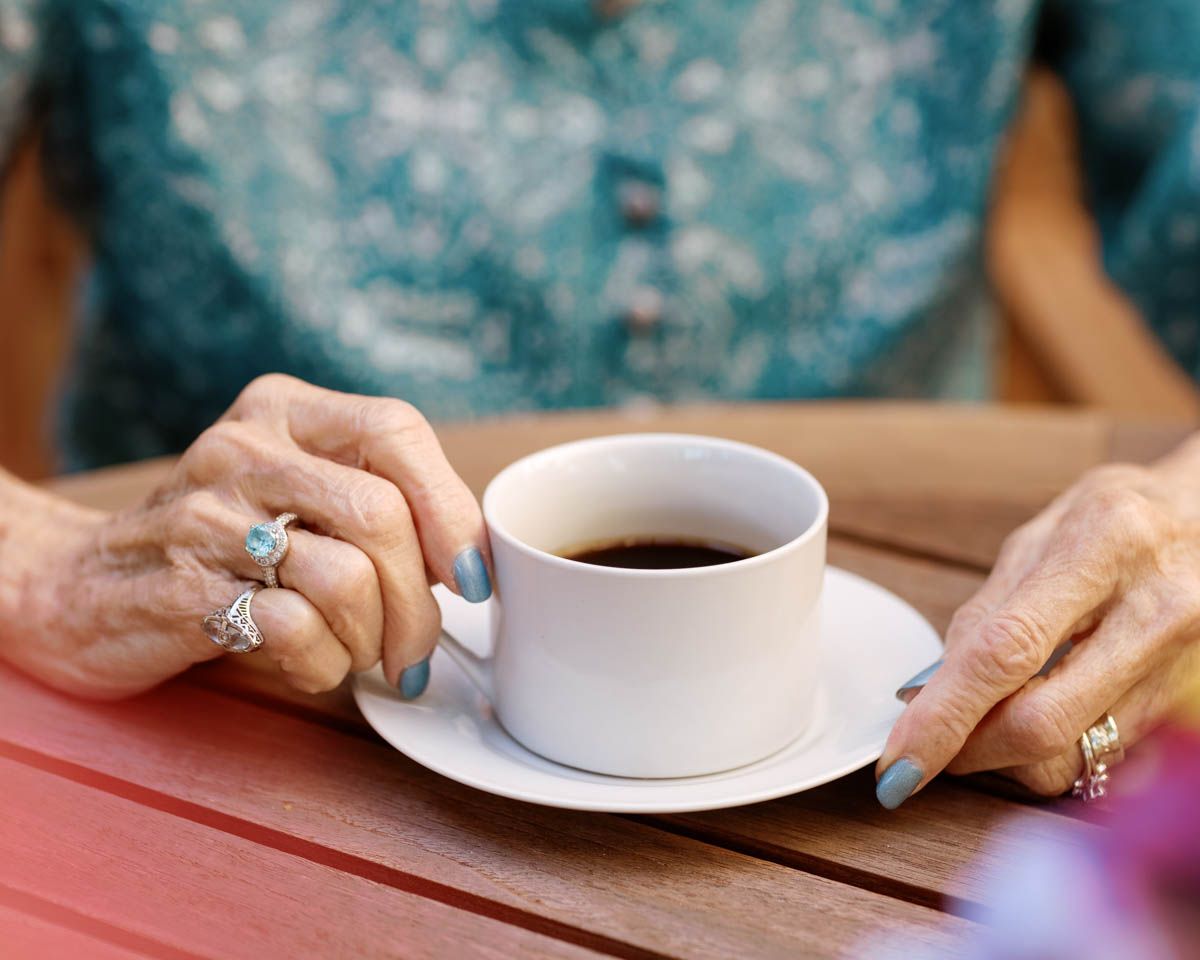 Close-up of elderly woman's hands holding a white coffee cup on a red table.