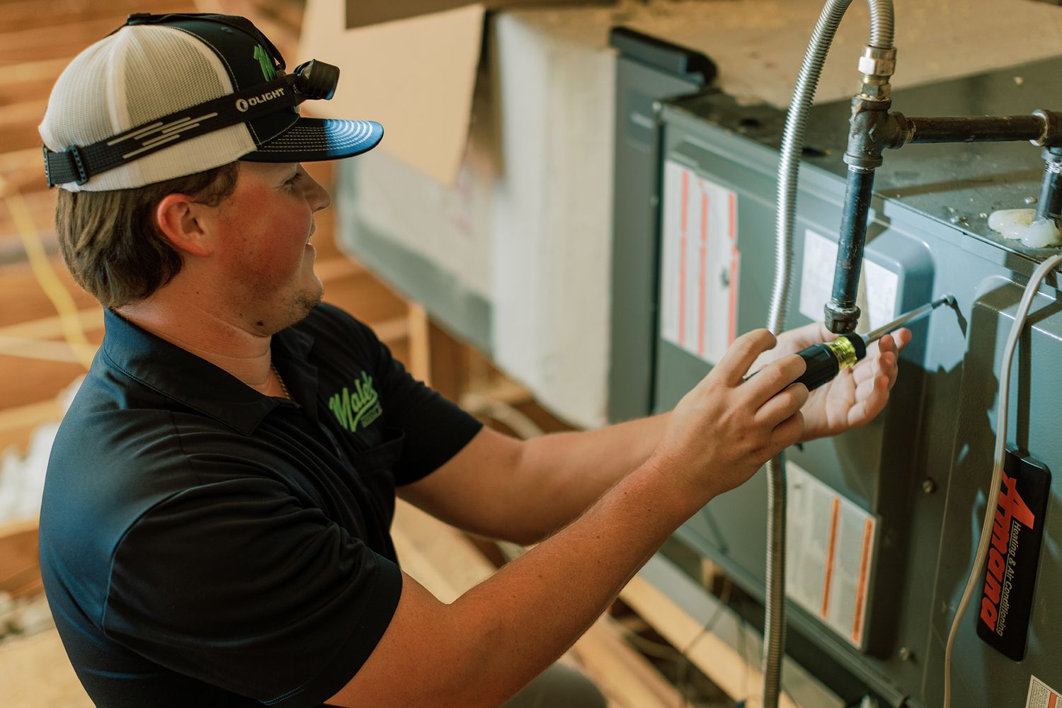 HVAC technician in black polo and cap installing or repairing a furnace unit at a residential construction site.