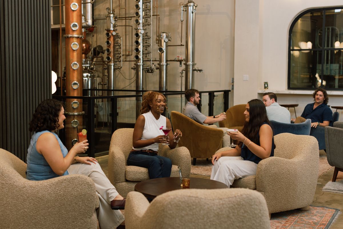 Group of friends relaxing with drinks in cozy lounge chairs inside a modern craft distillery with copper stills.