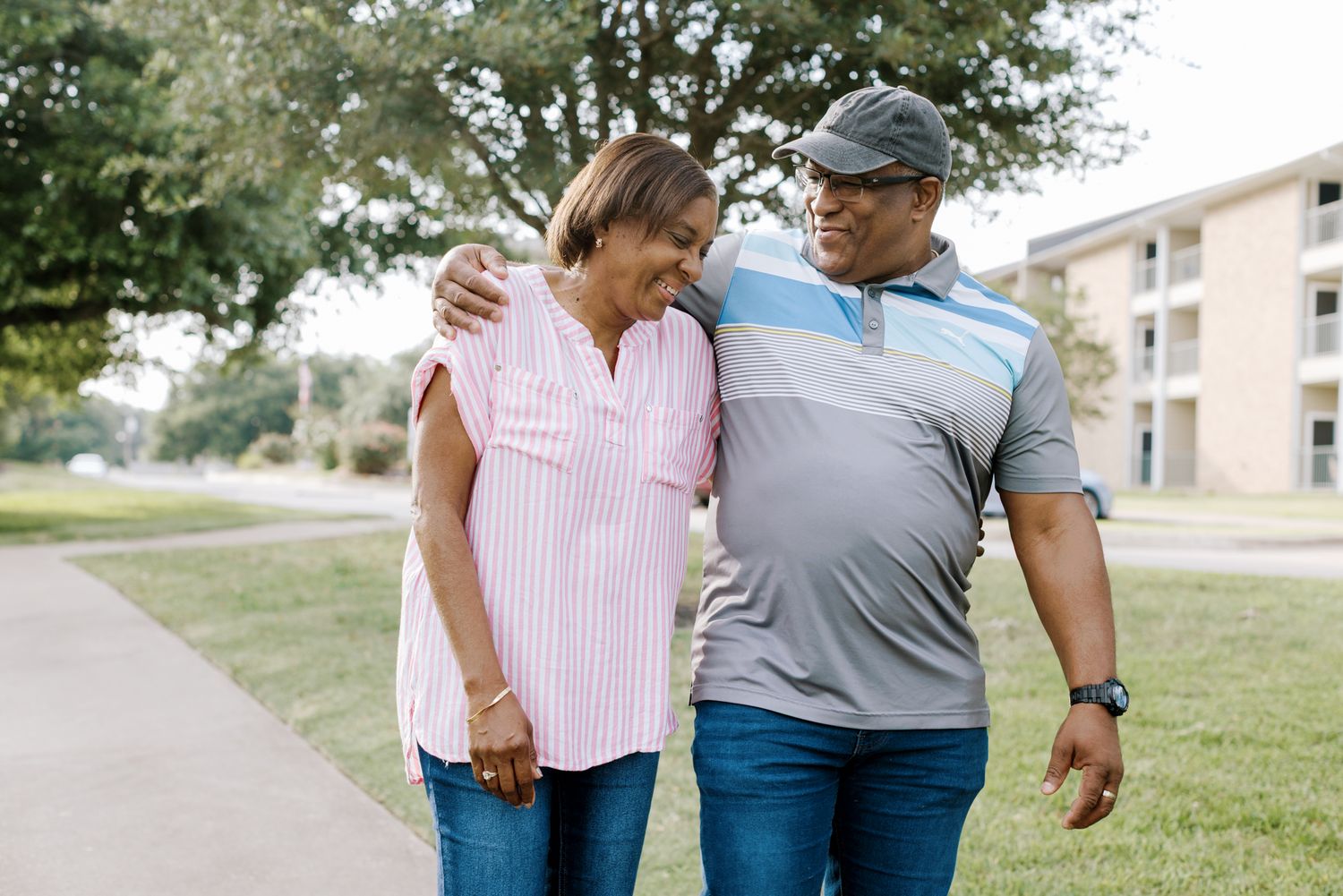 A smiling couple walks arm in arm on a sunny day near a green lawn and apartment building.