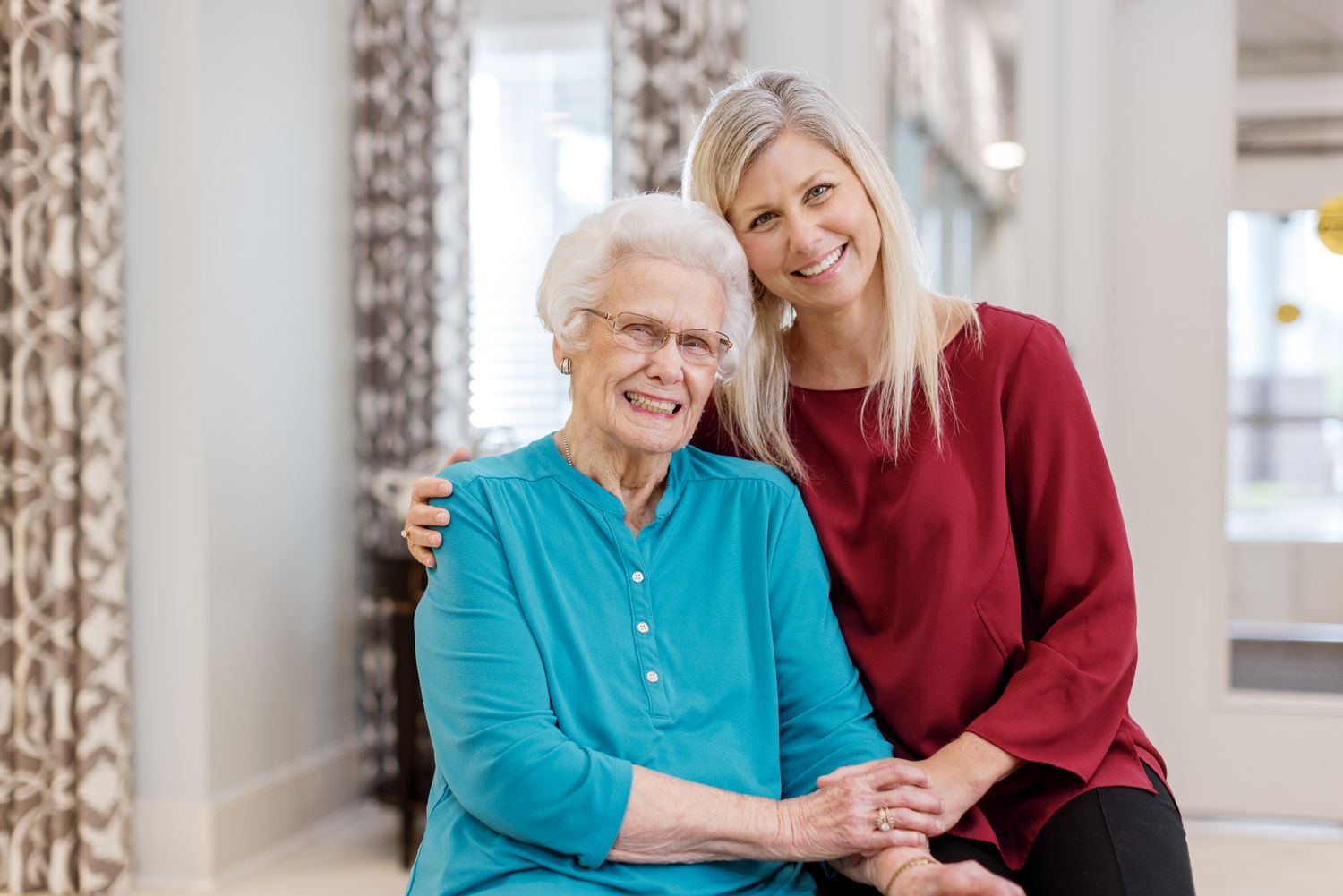Elderly woman and young blonde caregiver smiling together, holding hands in a bright, welcoming senior living community.