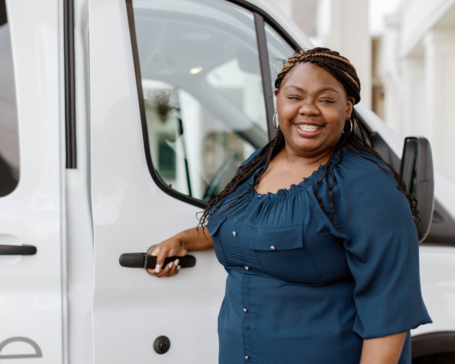 A smiling Black woman in a blue dress stands beside a white van, holding keys in a sunny parking lot.