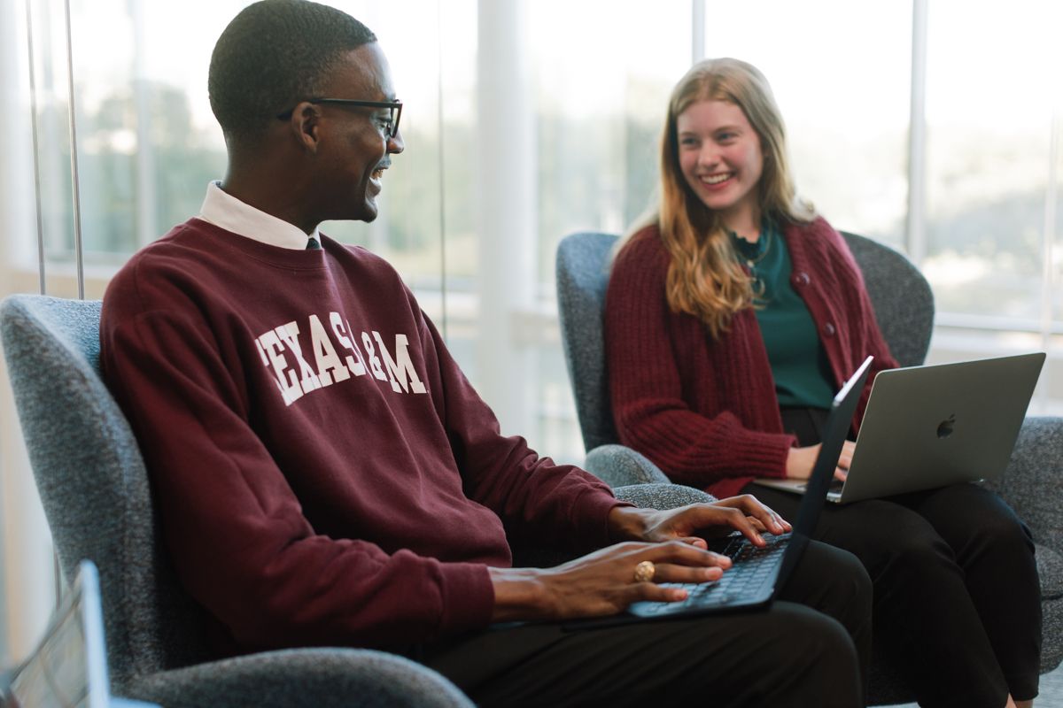 Two Texas A&M students work on laptops in a bright modern campus lounge area.