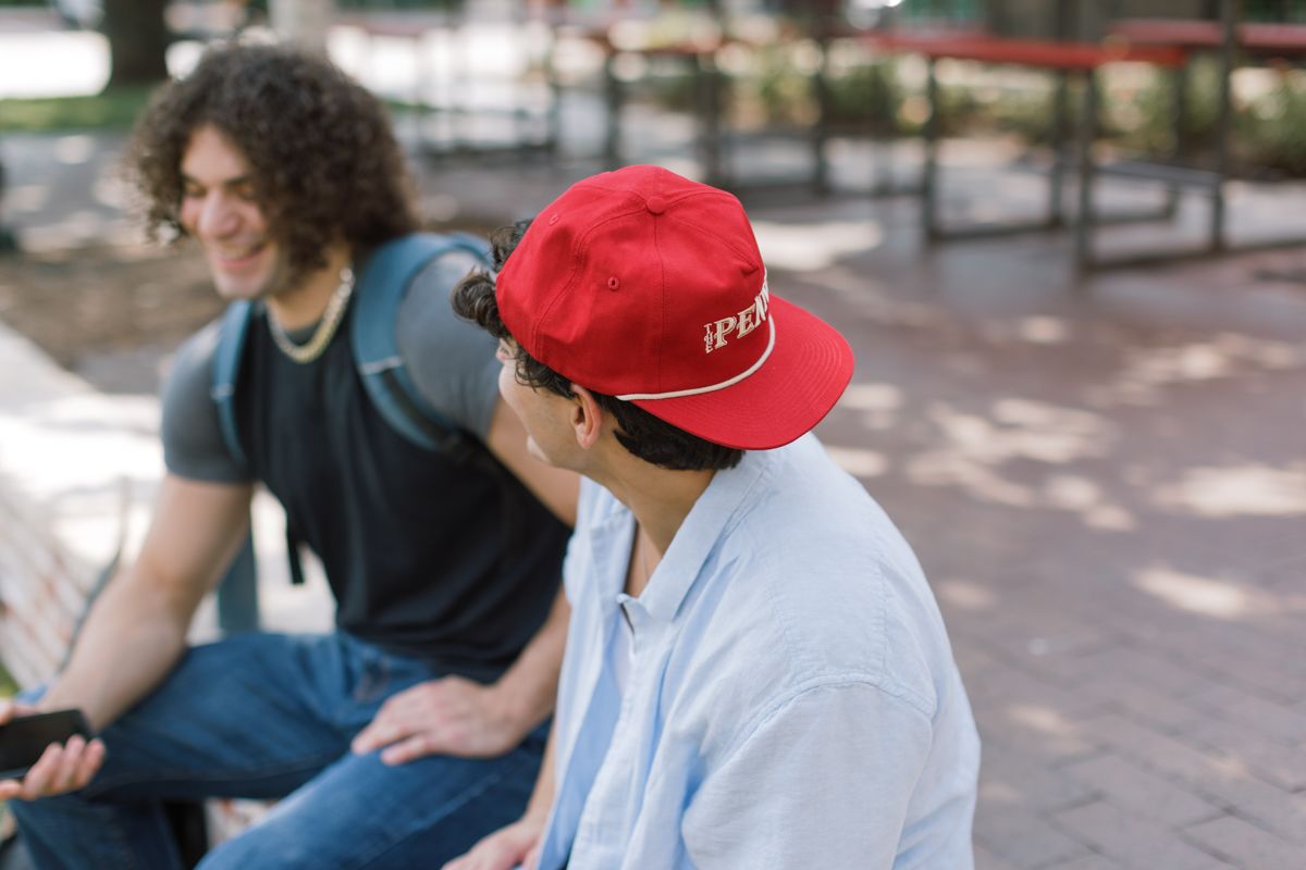 Two young men sit outdoors, one wearing a red cap, chatting in a park setting.