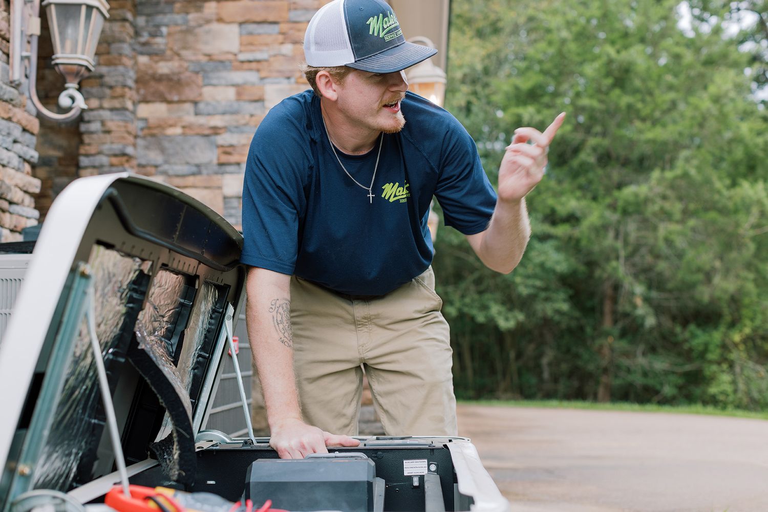 Technician in navy shirt and cap gestures while working on golf cart engine with toolbox outdoors.