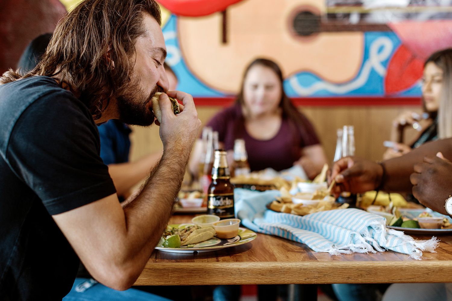 Man with long hair eats at a restaurant table with friends, food, and beer bottles in colorful setting.