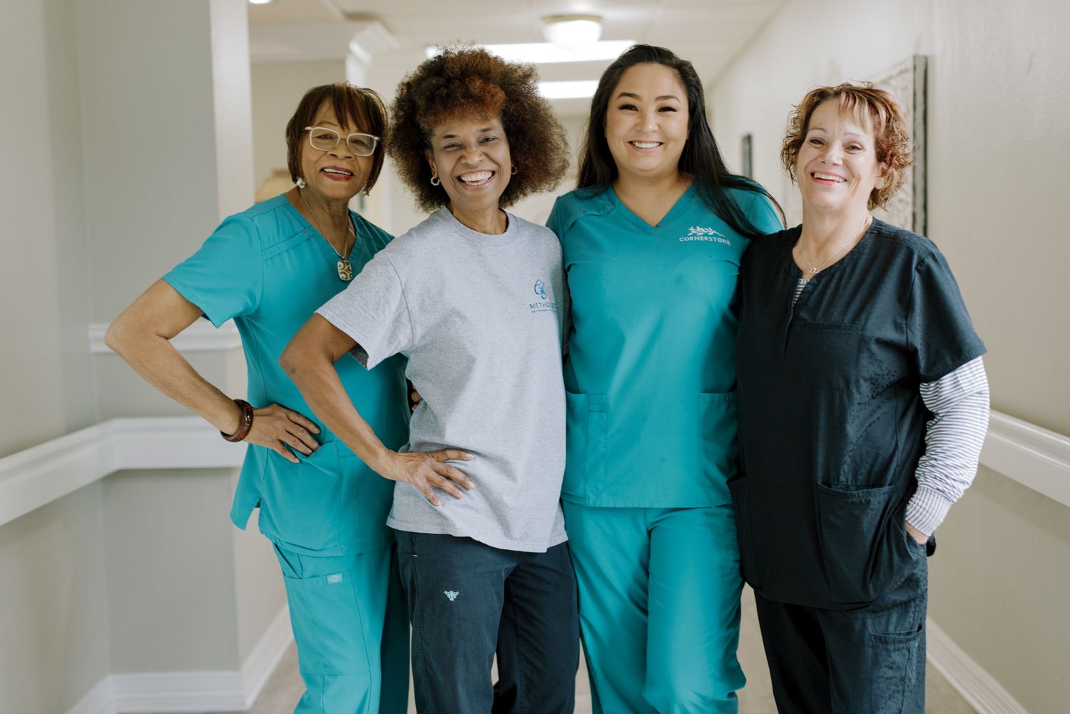 Four female healthcare workers in teal and dark scrubs pose together smiling in a bright hospital hallway.