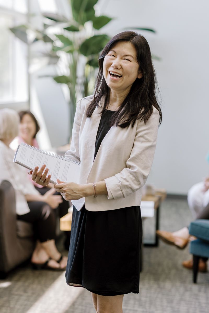 A smiling Asian businesswoman in a blazer holds a notebook while presenting to colleagues in a bright modern office.