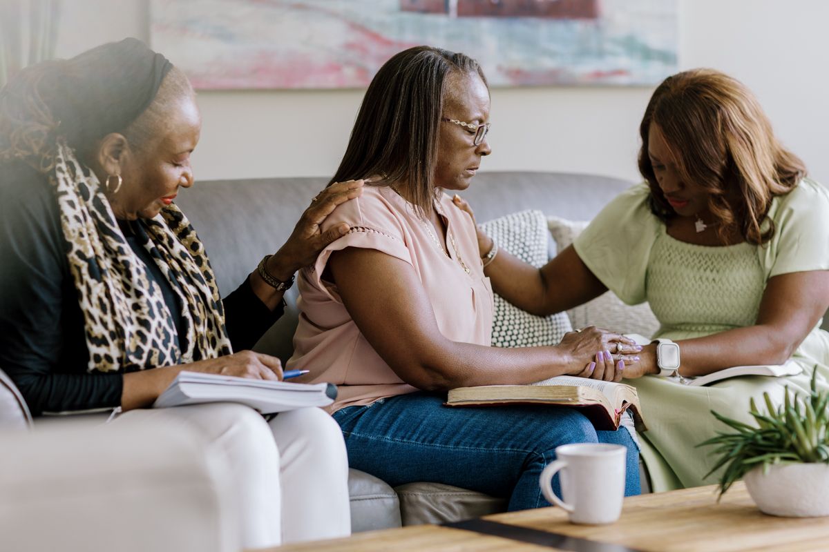Three women sitting together on a couch, praying and reading the Bible in a cozy living room setting.