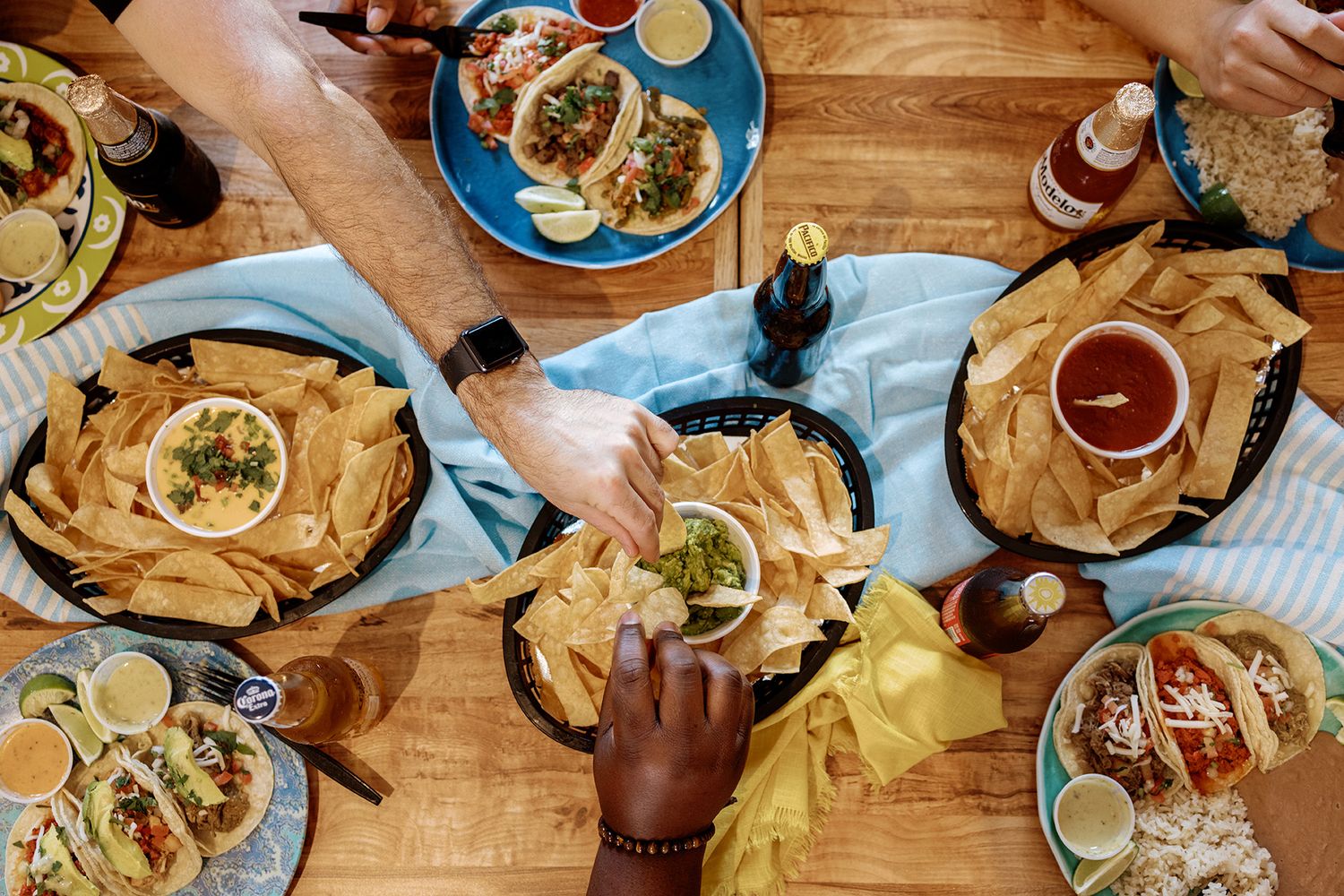 Overhead view of friends sharing Mexican food with nachos, tacos, and drinks on a wooden table.