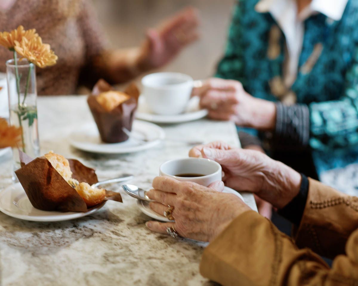 Two elderly women enjoying coffee and muffins together at a cafe table in a warm, cozy setting.