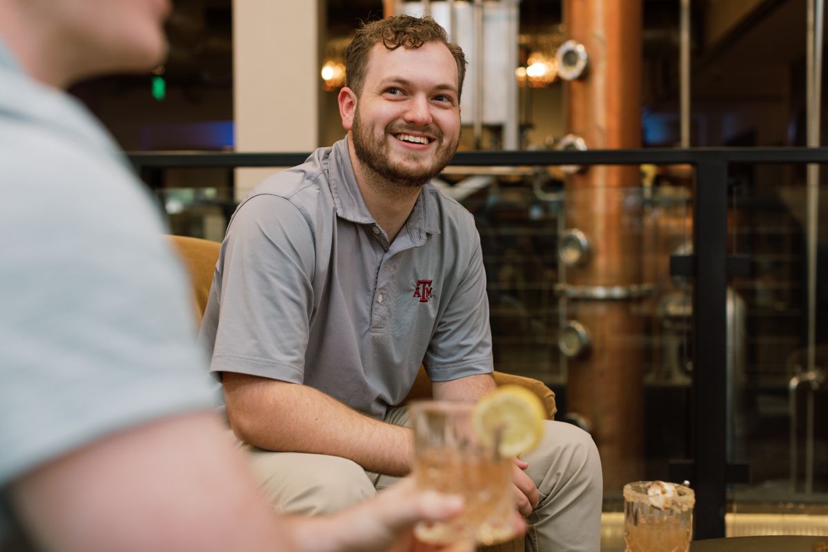 Smiling man in grey polo shirt enjoying a whiskey cocktail at a distillery bar with copper stills behind him.