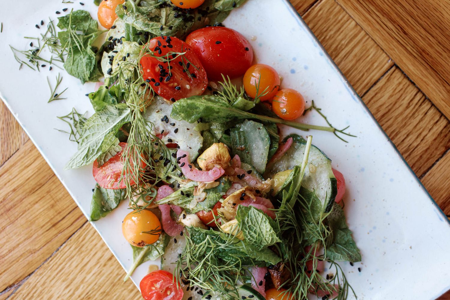 Fresh heirloom tomato salad with arugula, dill, and mixed greens on a white rectangular plate.