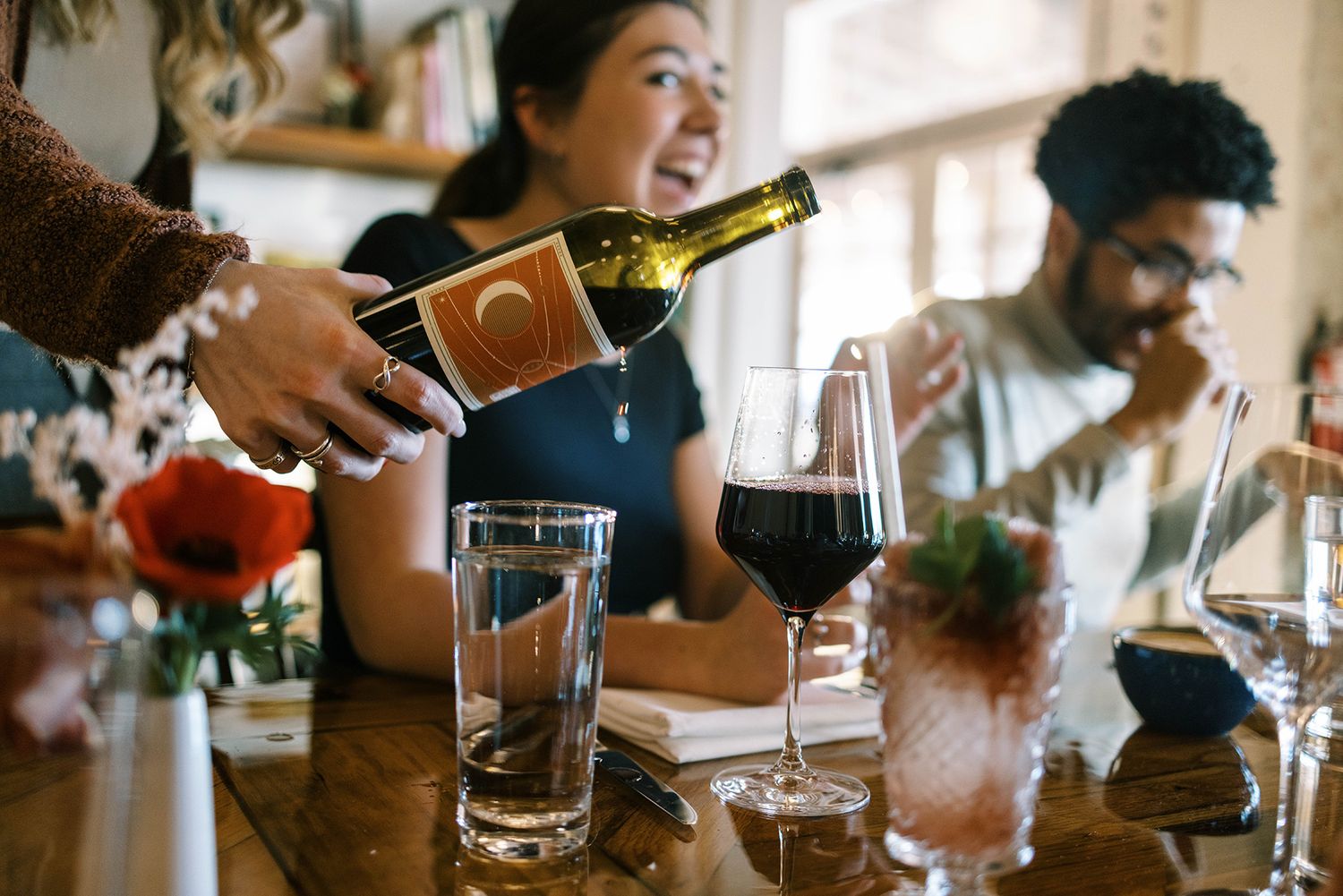 Server pouring red wine into glass at restaurant table with smiling guests enjoying drinks and conversation.