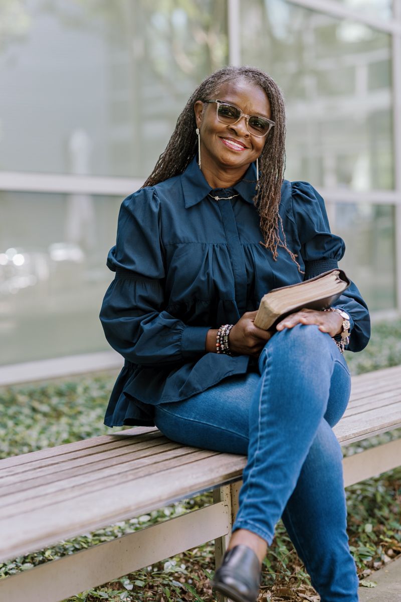 Smiling woman with braids and glasses sits on bench holding a book, wearing navy blouse and jeans outdoors.