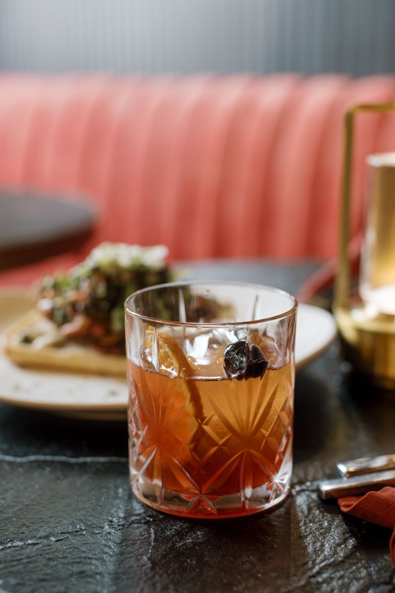 Amber cocktail in crystal glass with cherry garnish on dark table, blurred pink booth and appetizer plate in background.