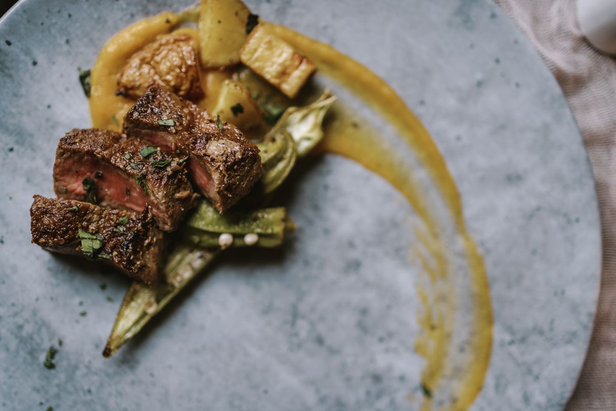 Gourmet seared beef with roasted vegetables and golden sauce on a rustic blue ceramic plate.