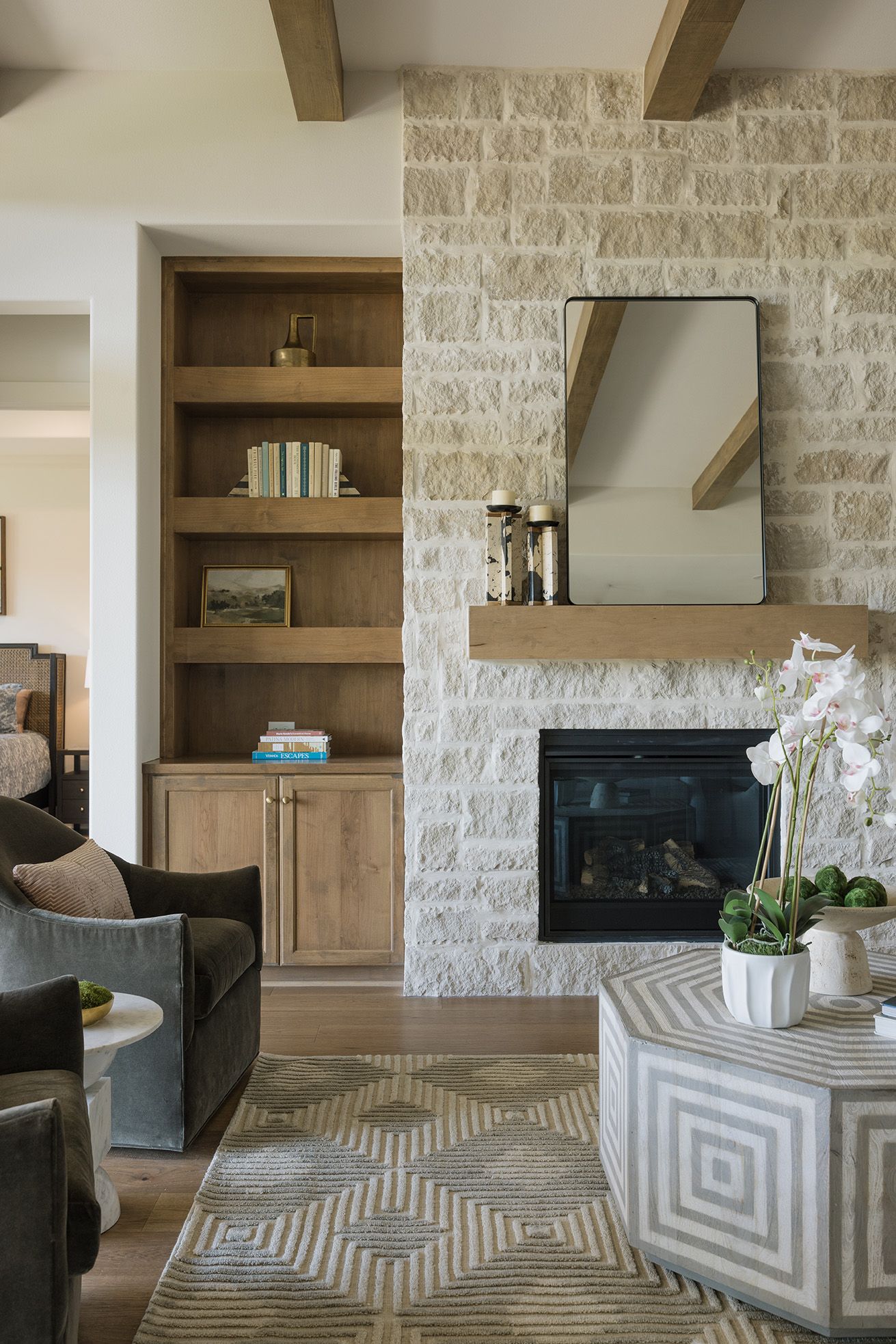 Cozy living room with limestone fireplace, built-in wood shelving, exposed beams, and geometric patterned rug.