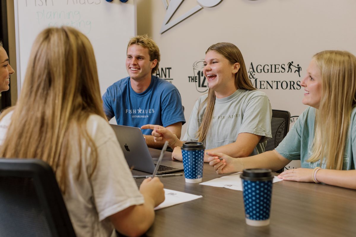 Group of college students laughing together around a conference table with Texas A&M branded cups and laptops.