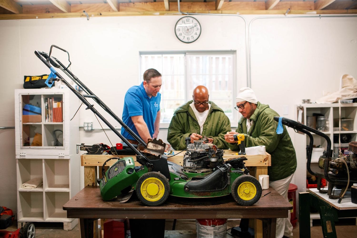 Three men repairing a John Deere lawn mower on a workbench in a workshop.